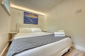 A hotel room with a bed, a painting, a nightstand, and a wall-mounted light.