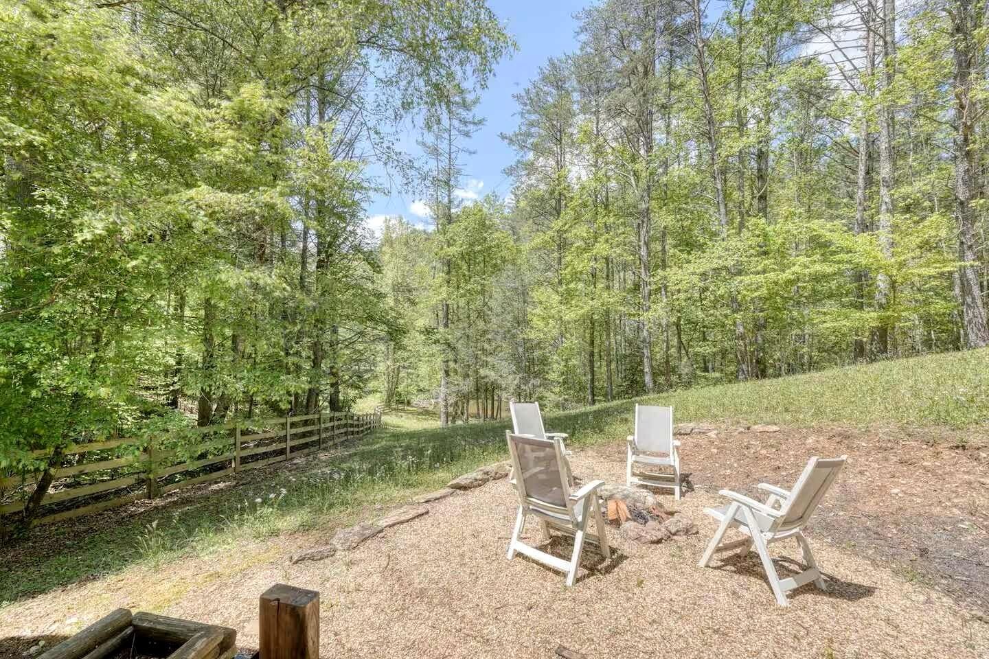 A forested area with a gravel path, two white chairs, and a small fire pit.