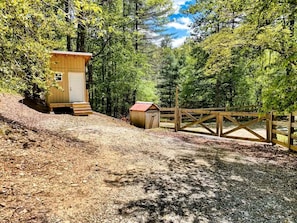 A small wooden cabin with a door and steps, surrounded by a forest and a gravel path.