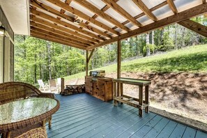 Une terrasse extérieure couverte, dotée d’une table et de chaises, d’un meuble en bois et offrant une vue sur une colline boisée.
