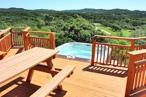 A wooden deck with a hot tub, picnic table, and wooden railings overlooking a green landscape.