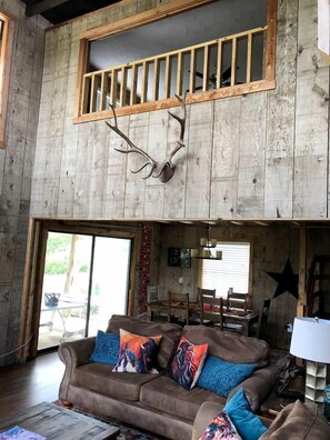 Un intérieur rustique avec un plafond en bois, une tête de cerf accrochée au mur, un canapé marron agrémenté de coussins colorés et un coin repas avec des chaises et une table.