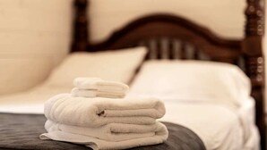 Una cama bien hecha con cabecera de madera, sábanas blancas y una pila de toallas dobladas.