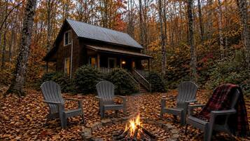 A cabin with a fire pit and three Adirondack chairs in a forest.
