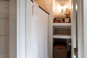 Un pasillo estrecho con una puerta blanca, una toalla blanca colgada en un perchero, una cesta de mimbre y una lámpara de pared.