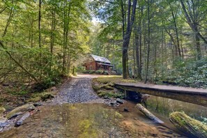 Una cabaña rústica rodeada por un bosque, un puente de piedra y un pequeño arroyo.
