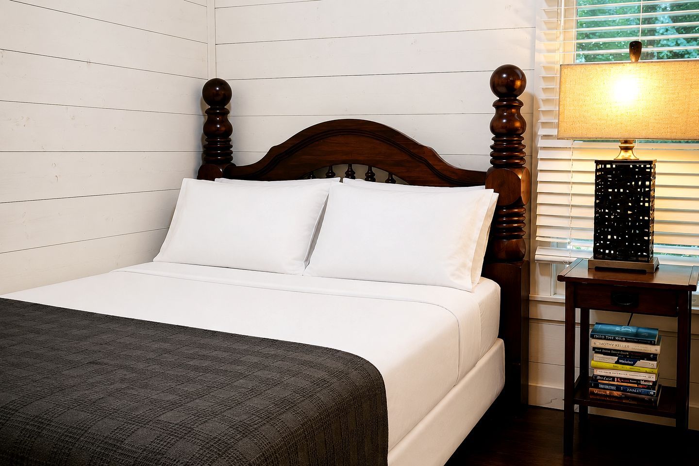 A bed with white bedding, a wooden headboard, and a bedside table with a lamp and books.