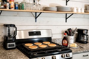 A kitchen with a stove, pancakes cooking, a coffee maker, and various kitchen items on shelves.