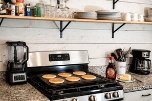 A kitchen with a stove, pancakes cooking, a coffee maker, and various kitchen items on shelves.
