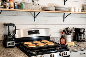 A kitchen with a stove, pancakes cooking, a coffee maker, and various kitchen items on shelves. - Secluded Cabin on Suches Creek and in the National Forest for a Relaxing Georgia Getaway (Suches)