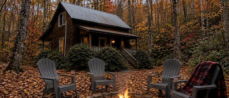Una cabaña con un brasero y tres sillas Adirondack en un bosque.