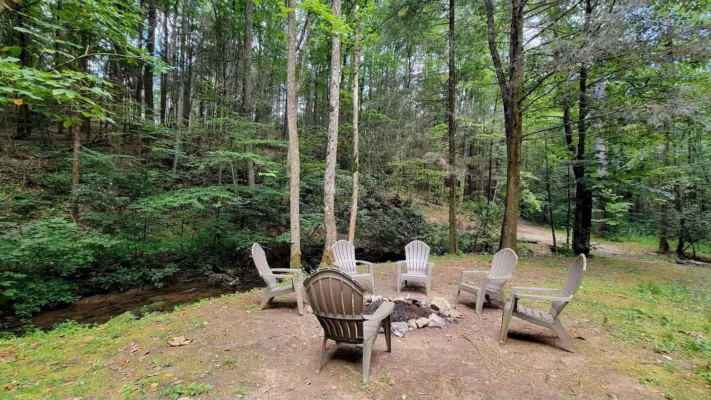 A forest setting with a fire pit and Adirondack chairs.