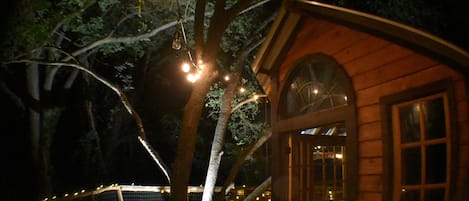 Una cabaña de madera con un amplio ventanal arqueado, rodeada de árboles e iluminada por luces colgantes.