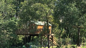 Une cabane en bois perchée dans un arbre, accessible par un escalier, et entourée d’un feuillage dense.