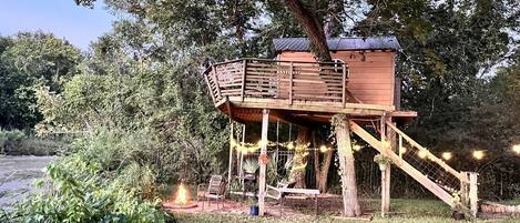 Una casa en el árbol de madera con una plataforma y escaleras, rodeada de vegetación y una masa de agua.
