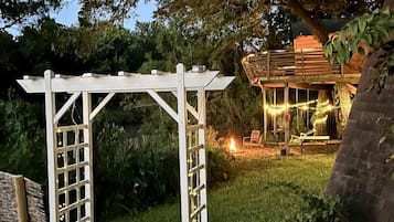 A white pergola structure in a garden setting with a wooden building in the background.