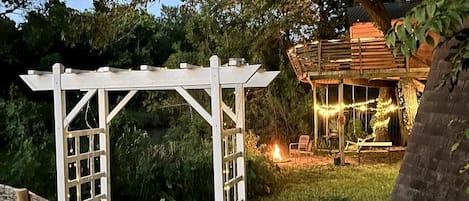 Une pergola blanche, installée dans un jardin, avec un bâtiment en bois en arrière-plan.