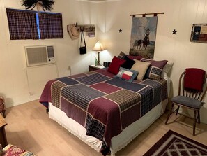 Un dormitorio con una cama grande, una mesita de noche con una lámpara, una silla, una ventana con cortinas, un calentador de pared y un cuadro con temática vaquera.