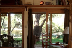 A wooden interior with a view of a lake and trees, a chair, and a table with a plate and a book. - Pet-Friendly Cabin Rental with Creekside Views near Bandera, Texas (Pipe Creek)
