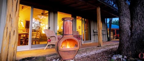A rustic cabin with a large fire pit in the foreground, wooden beams, and a covered patio area with outdoor seating.