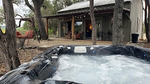 A hot tub with a rustic wooden deck, a covered seating area, and a view of a cabin. - Pet-Friendly Cabin Rental with Creekside Views near Bandera, Texas (Pipe Creek)