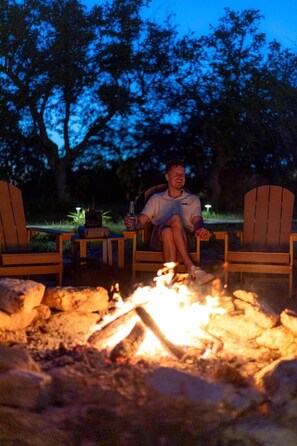 A person sitting on a wooden chair by a campfire with a bottle in hand. - A Romantic Getaway in this Luxurious Yurt for Couples in Wimberley, TX (Wimberley)