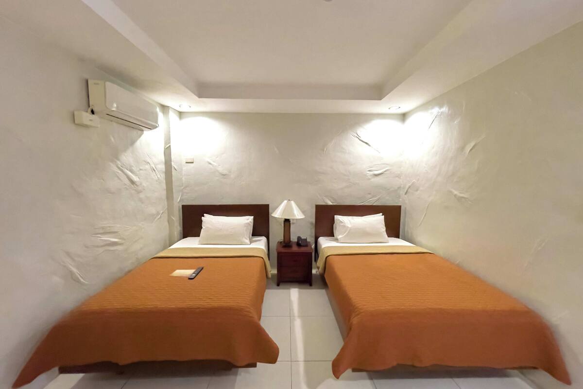 Una habitación de hotel con dos camas individuales, una mesita de noche con lámpara y un equipo de aire acondicionado en la pared.