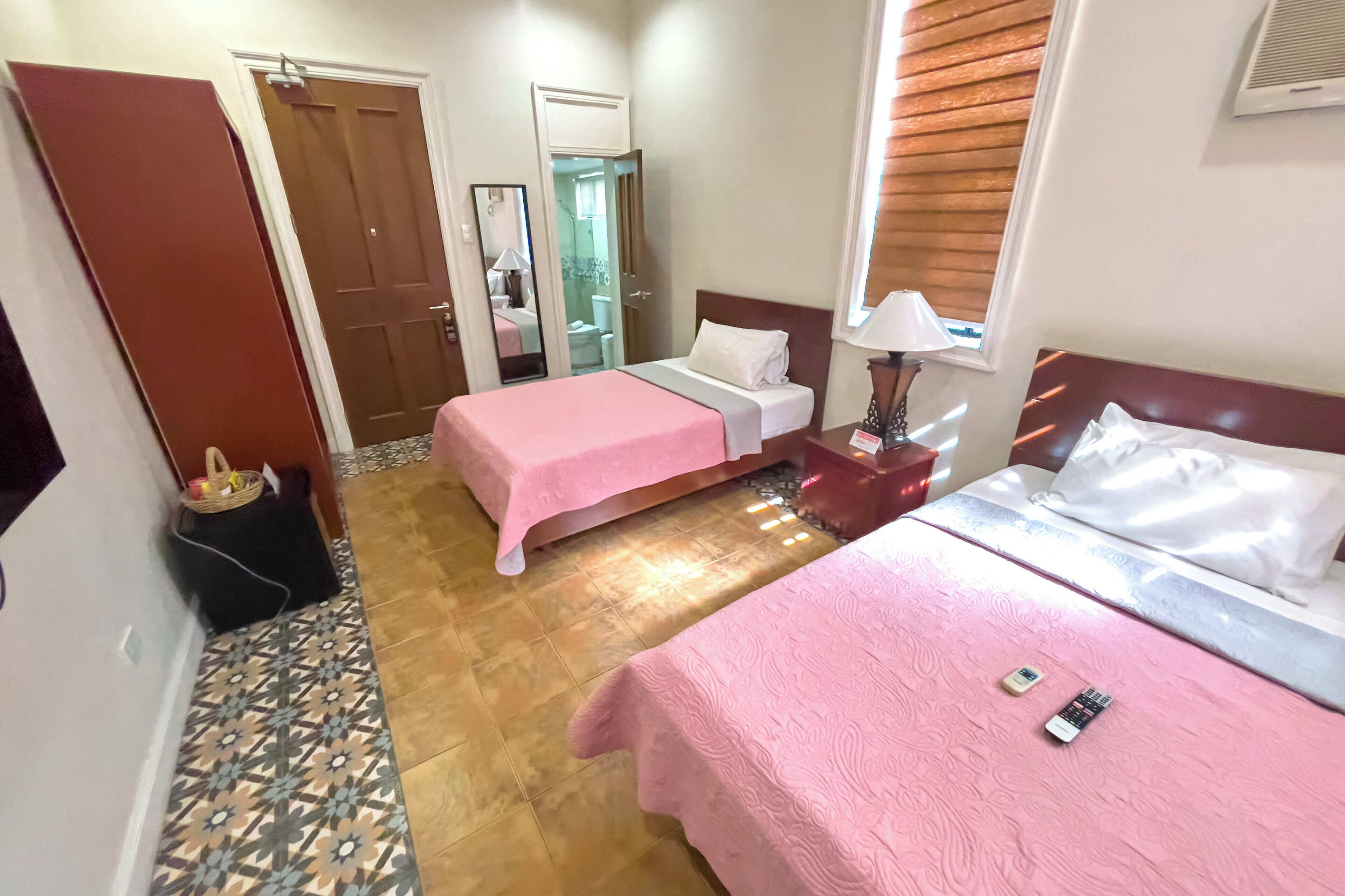 Habitación de hotel con dos camas, un espejo, una mesita de noche con lámpara y una ventana con persianas.