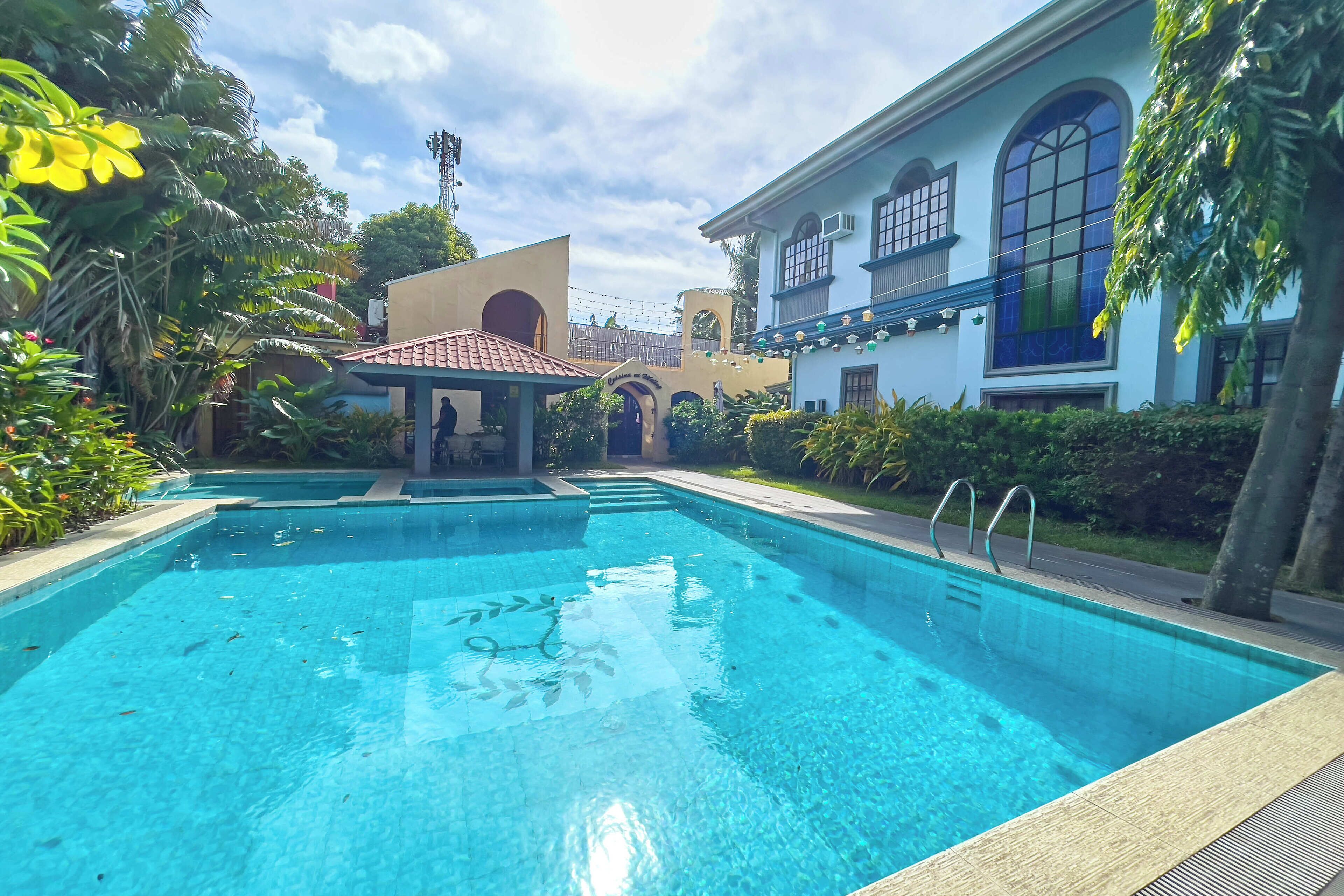 A clear swimming pool with a tiled deck, surrounded by lush greenery and a building with arched windows and a tiled roof.