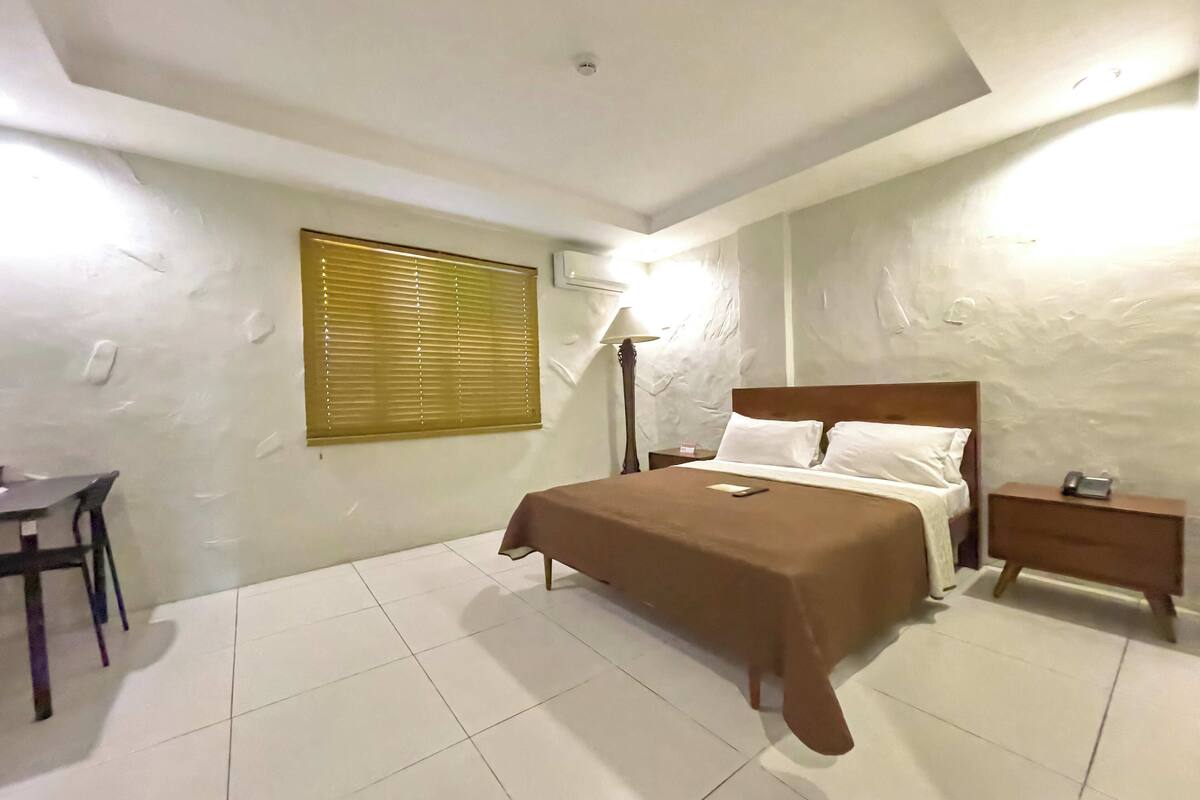 Un dormitorio con cama, mesita de noche, lámpara y una ventana con persianas.