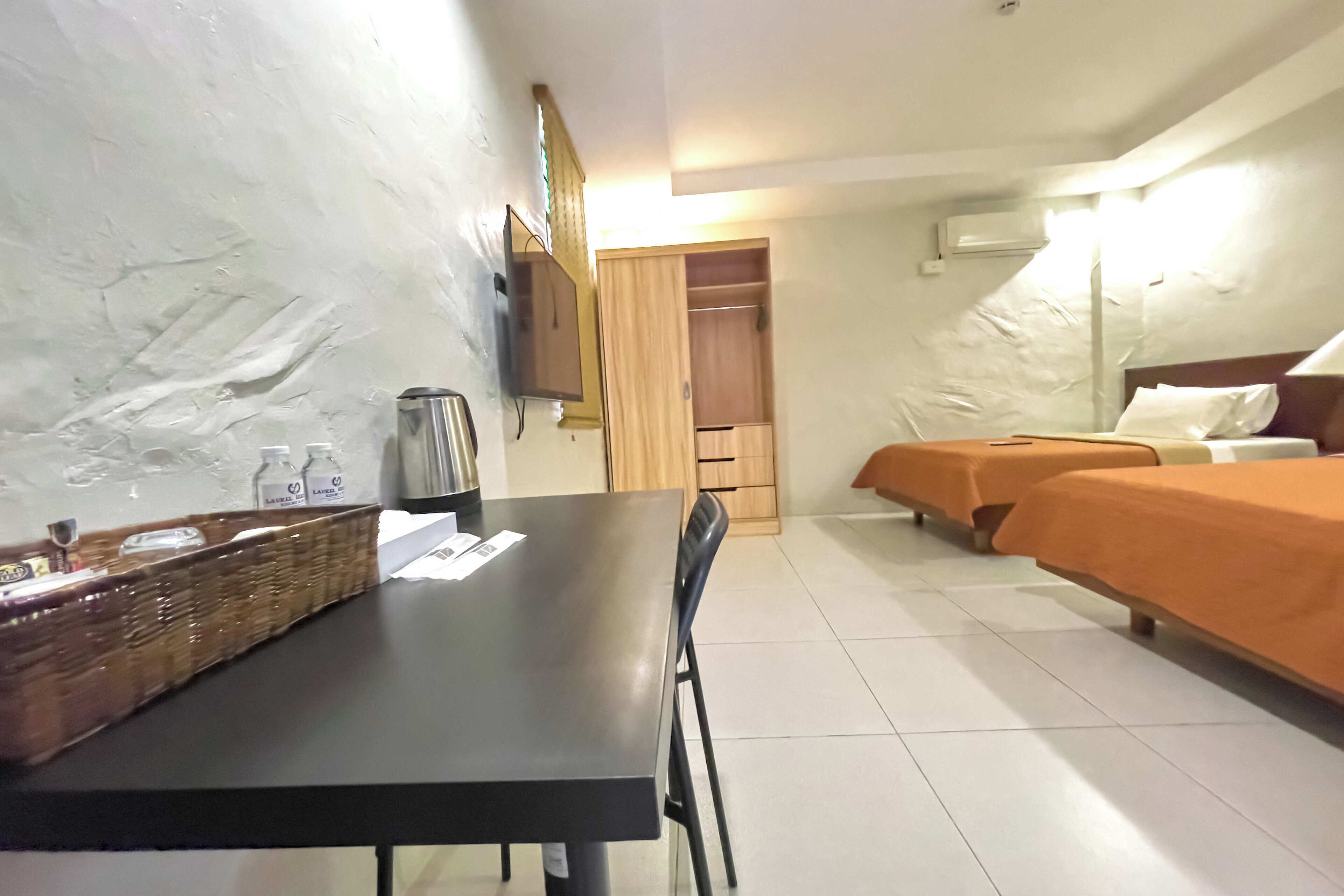 Habitació d'hotel amb taula de menjador, dos llits amb estovatges taronja, un armari de fusta i una televisió penjada a la paret.