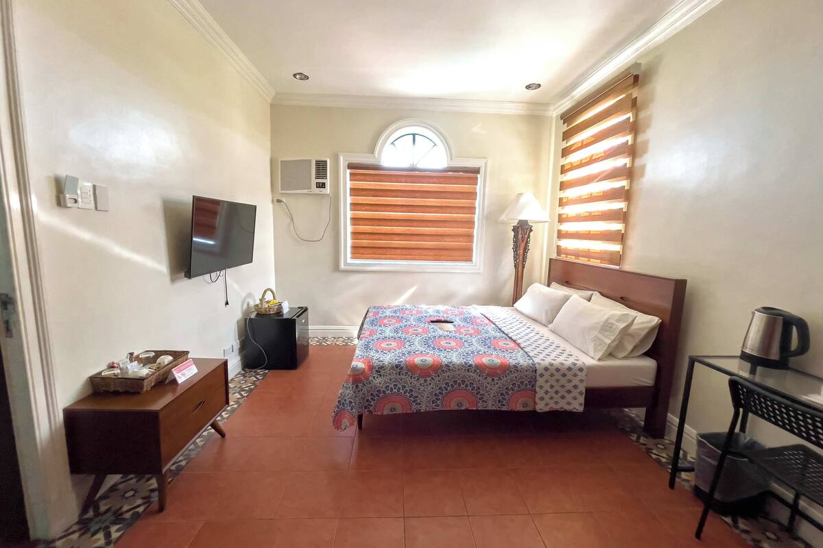Un dormitorio con cama, televisión, ventana con persianas, lámpara y una pequeña mesa con hervidor.