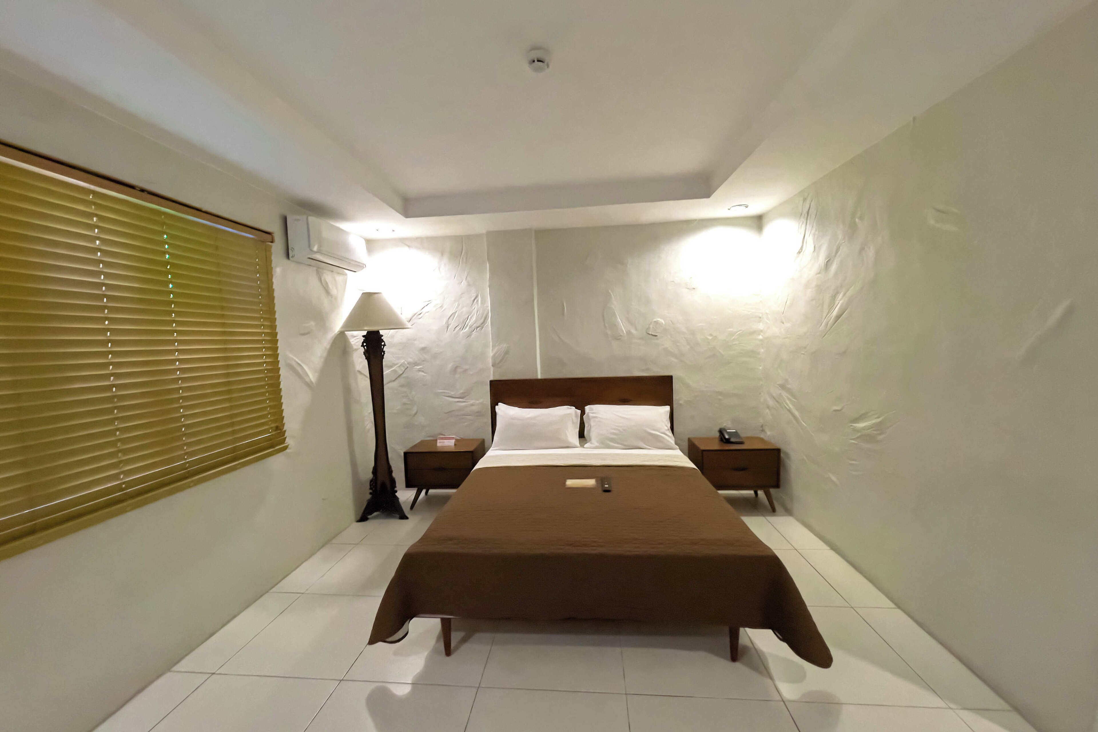A bedroom with a large bed, two bedside tables, a lamp, and a window with blinds.