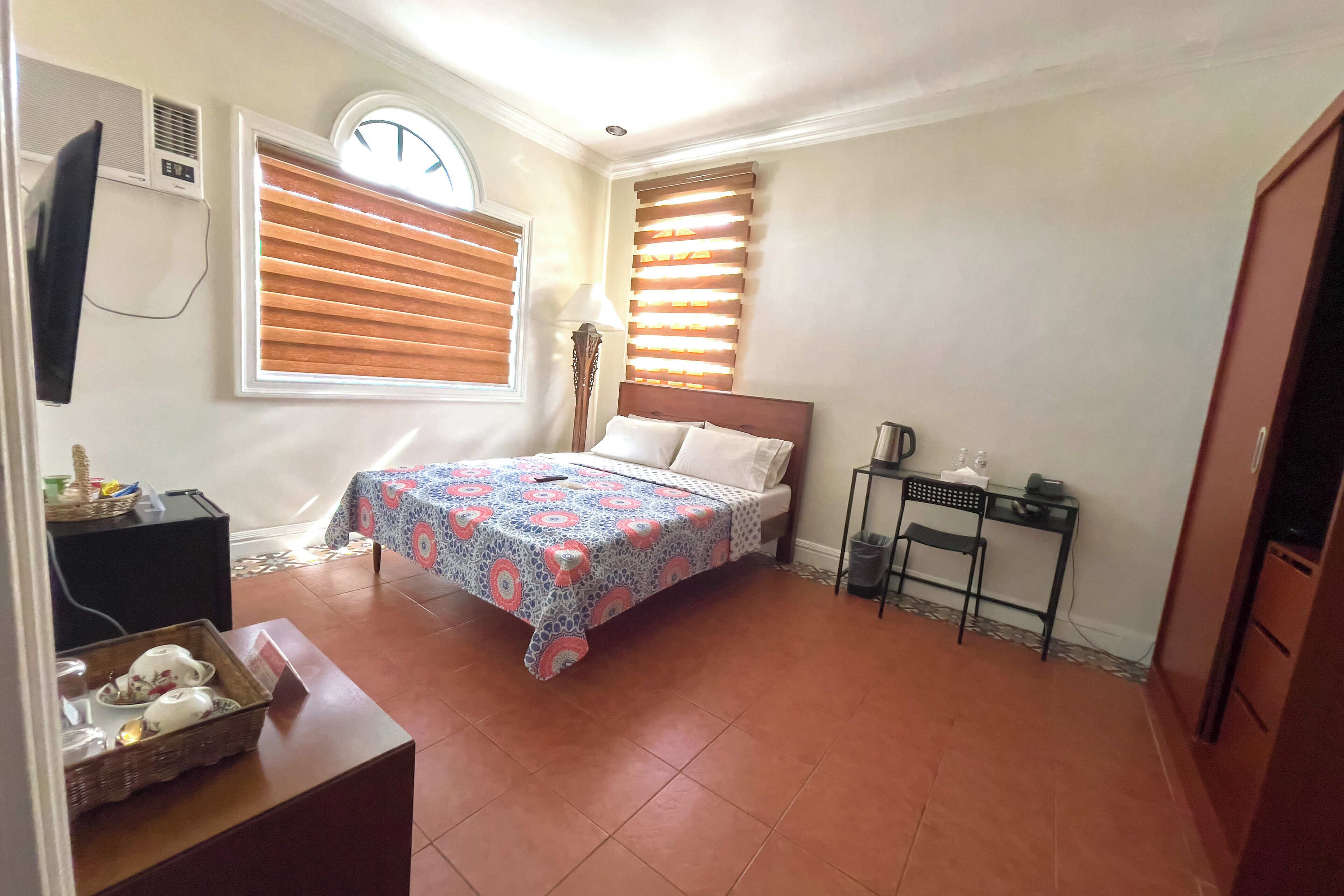 Une chambre à coucher avec un lit, une petite table, une chaise, une lampe et une fenêtre avec des stores.