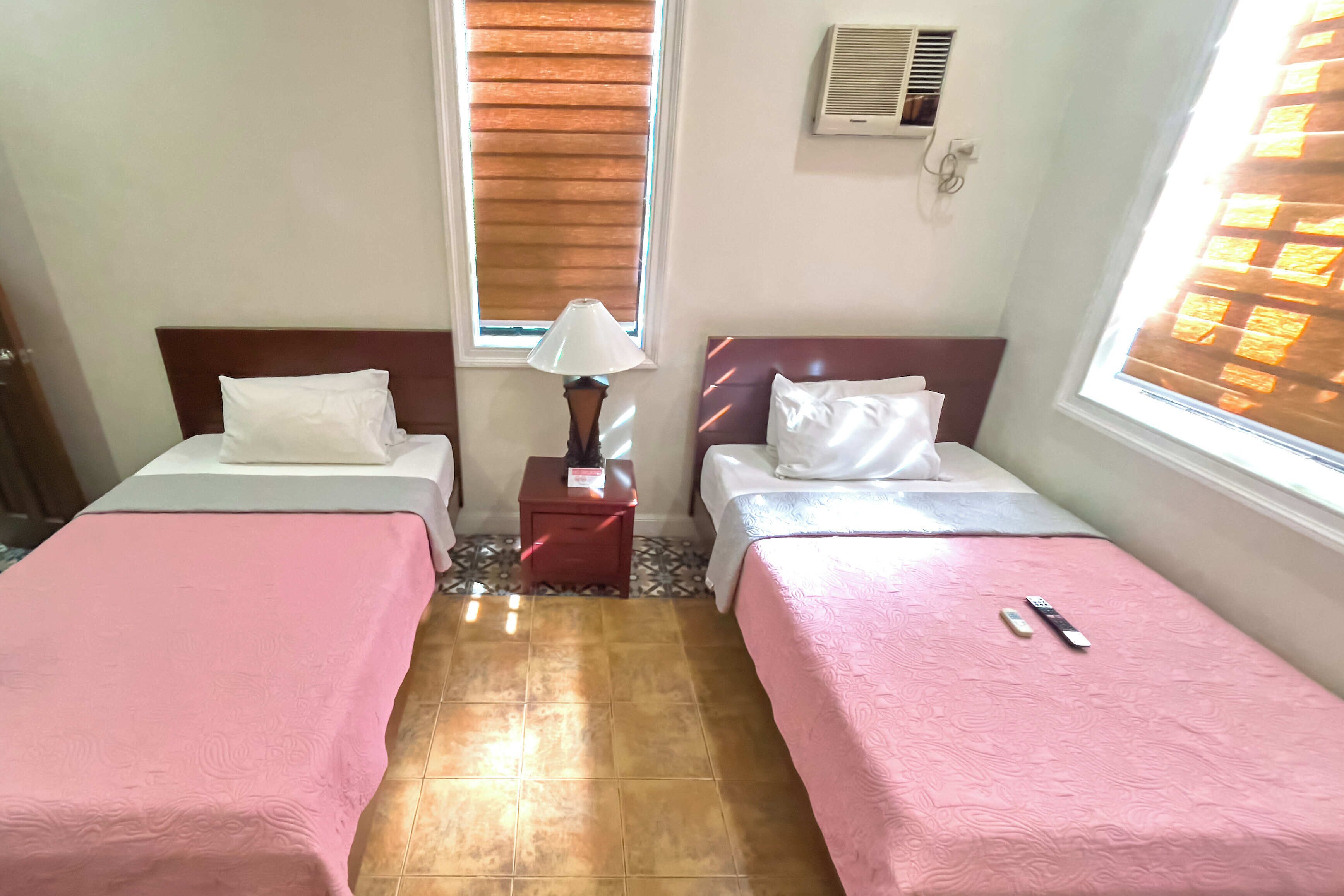 Una habitación con dos camas, cada una con una colcha rosa, una mesita de noche con lámpara y una ventana con persianas.