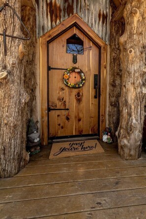 A wooden door with a wreath, a doormat with 'you're here!' text, and a rustic interior. - Hobbit Treehouse with Waterfall on the Brazos River for Glamping in Texas (Weatherford)