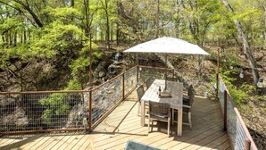 Eine Holzdielen-Terrasse mit Tisch und Stühlen, umgeben von einem Metallzaun, mit Blick auf ein bewaldetes Gebiet.