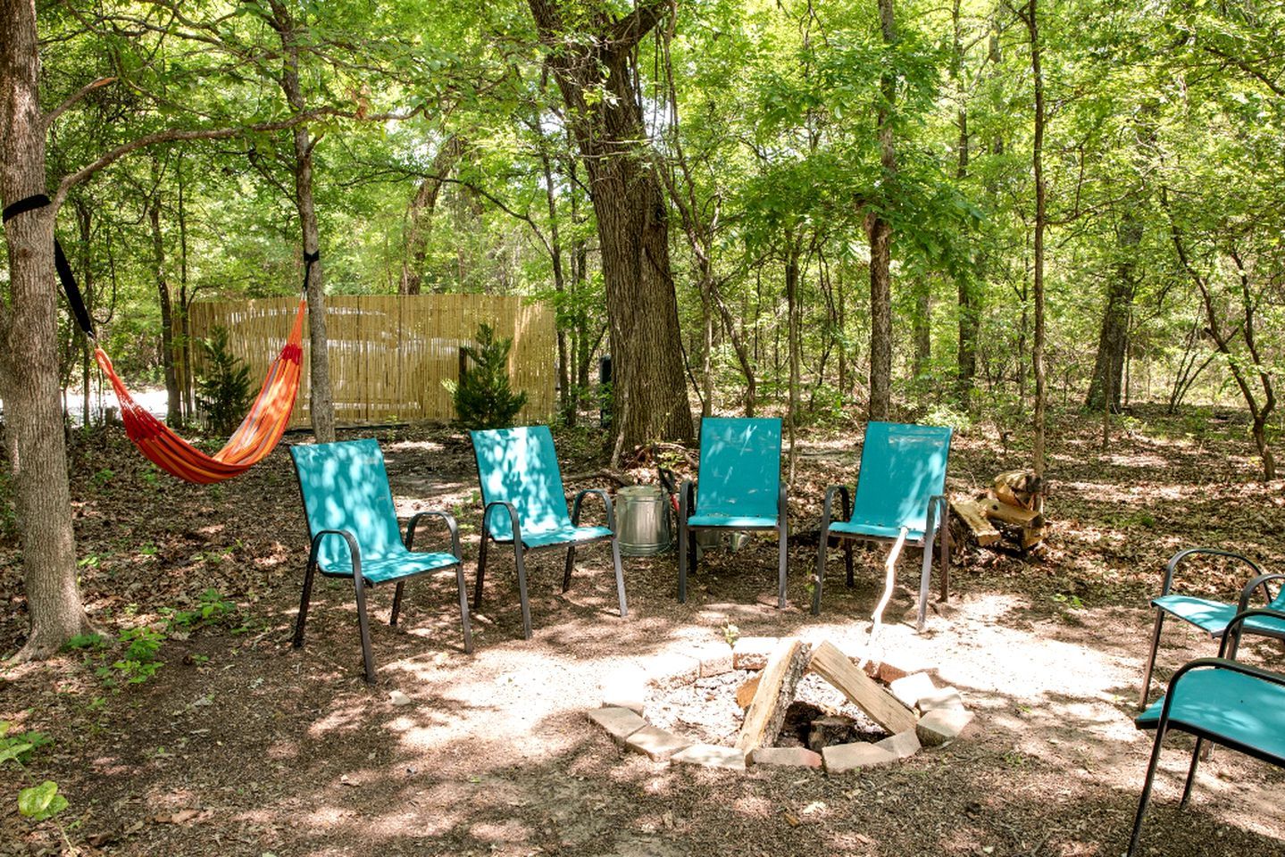 A forested area with a hammock, five green chairs, and a fire pit.