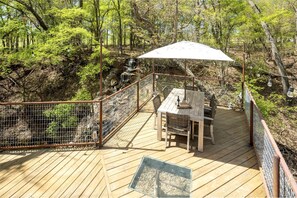 Une terrasse en bois, agrémentée d’une table et de chaises, entourée d’une clôture métallique, et offrant une vue sur une zone boisée.