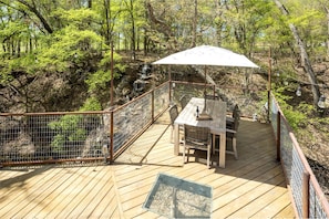 Une terrasse en bois, agrémentée d’une table et de chaises, entourée d’une clôture métallique, et offrant une vue sur une zone boisée.