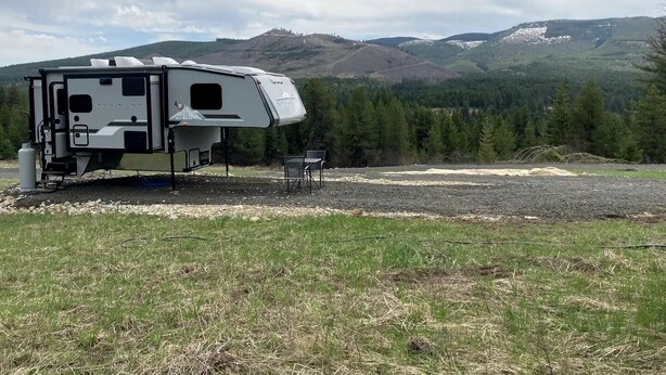 Una caravana blanca con zona de estar cubierta, una mesa pequeña y sillas, situada en un campo con montañas al fondo.