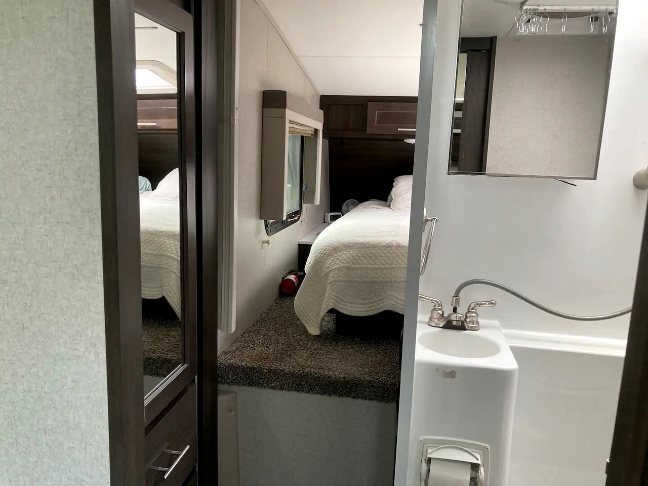 A compact bedroom with a single bed, a small window, a mirror, and a sink.