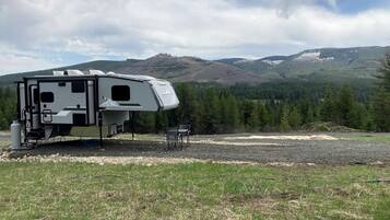 Una caravana blanca con zona de estar cubierta, una mesa pequeña y sillas, situada en un campo con montañas al fondo.