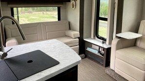 A kitchen area with a sink, a countertop, and a window. There is a seating area with a sofa and a chair. - Tiny Caravan Great for Couples with Landscape Views in Idaho (Saint Maries)