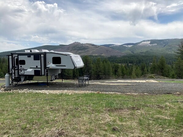 A white RV with a covered living area, a small table, and chairs, set in a grassy field with mountains in the background.