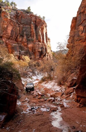 A rugged canyon with a jeep on a dirt path, surrounded by red rock formations and sparse vegetation.