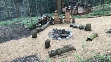 Un área boscosa con una fogata, dos sillas Adirondack y una terraza con mobiliario exterior.