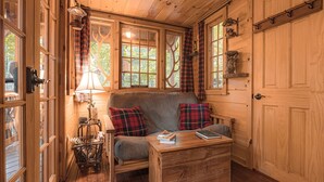 A cozy cabin interior with a sofa, a table with books, plaid curtains, and a wooden door. - Magical Tree House Rental in the Woods near Syracuse for Glamping in New York State (Lafayette)