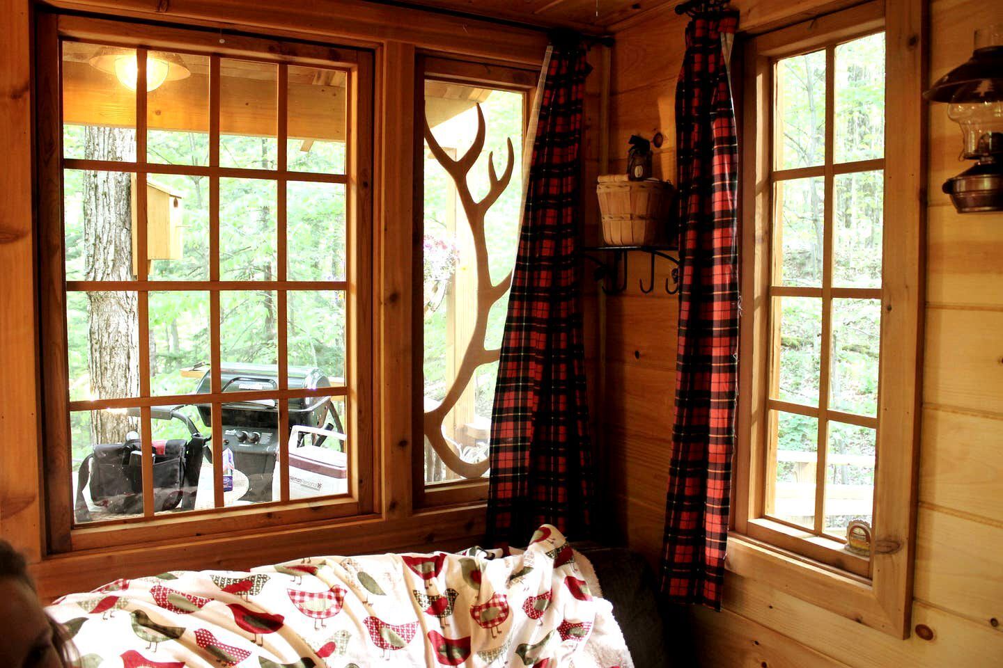 A cabin with large windows, a bed with a patterned blanket, plaid curtains, and a wooden interior.