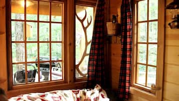 A cabin with large windows, a bed with a patterned blanket, plaid curtains, and a wooden interior.